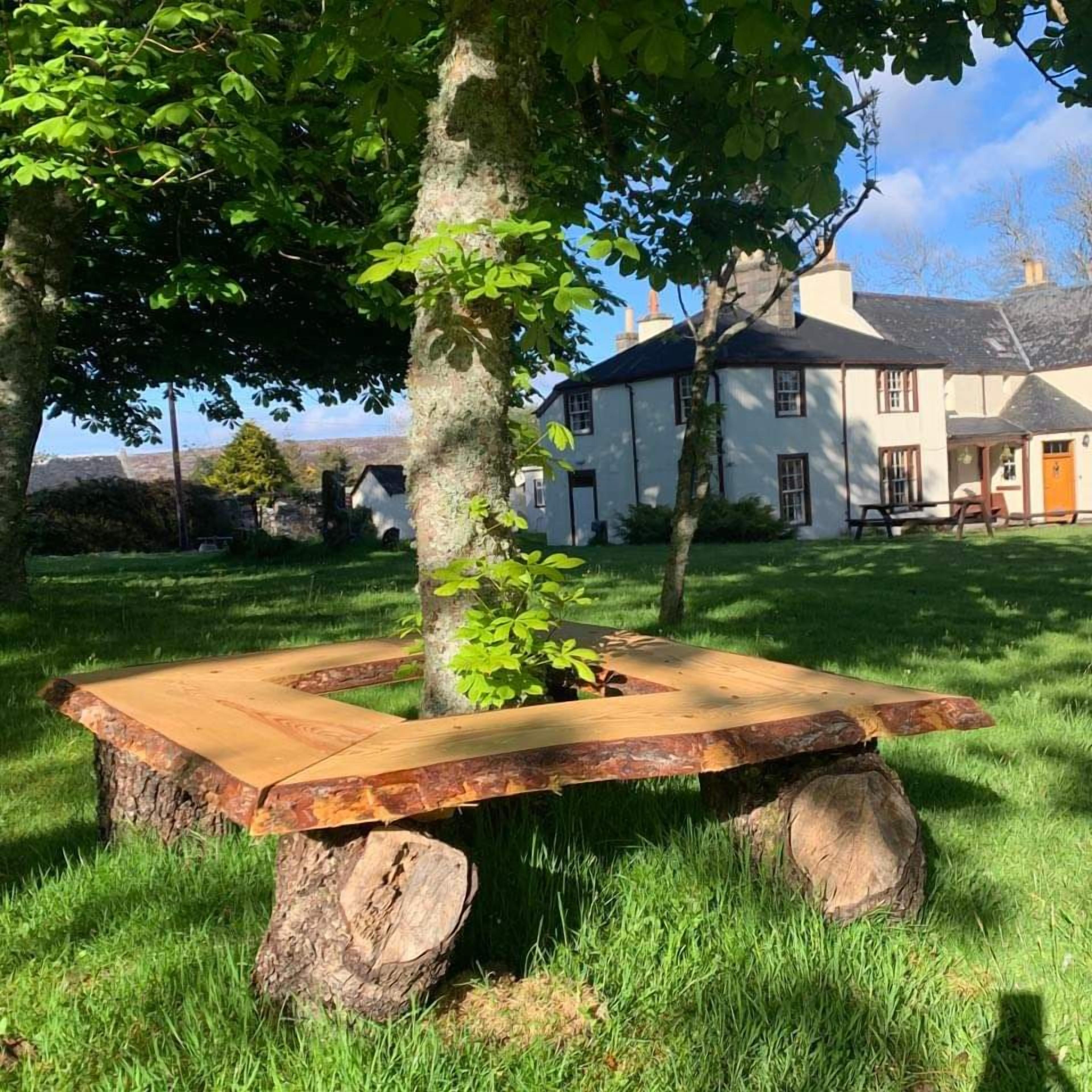 A rustic wooden bench with a tree trunk base, set in a grassy area with a white building in the background.