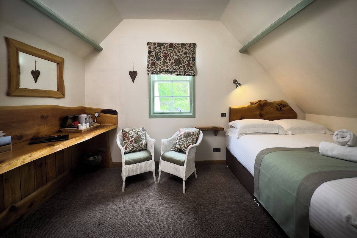 A cozy room with a bed, two chairs, a wooden bench, a window with a view, and a wall-mounted light fixture.
