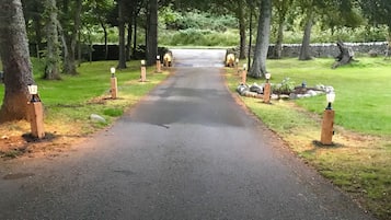 A paved pathway flanked by trees and wooden posts with lights, leading to a stone structure.