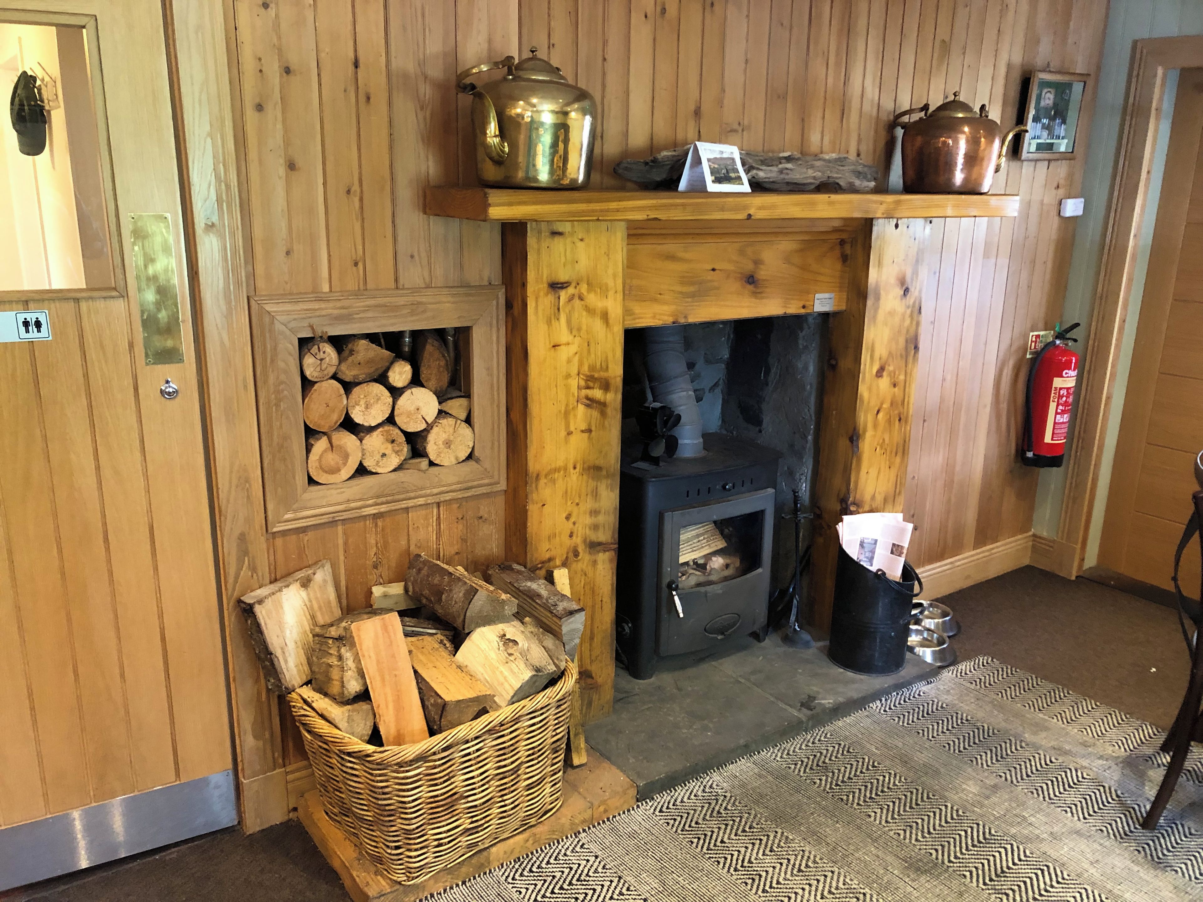 A wooden interior with a fireplace, a firewood storage area, and a metal kettle on a shelf.