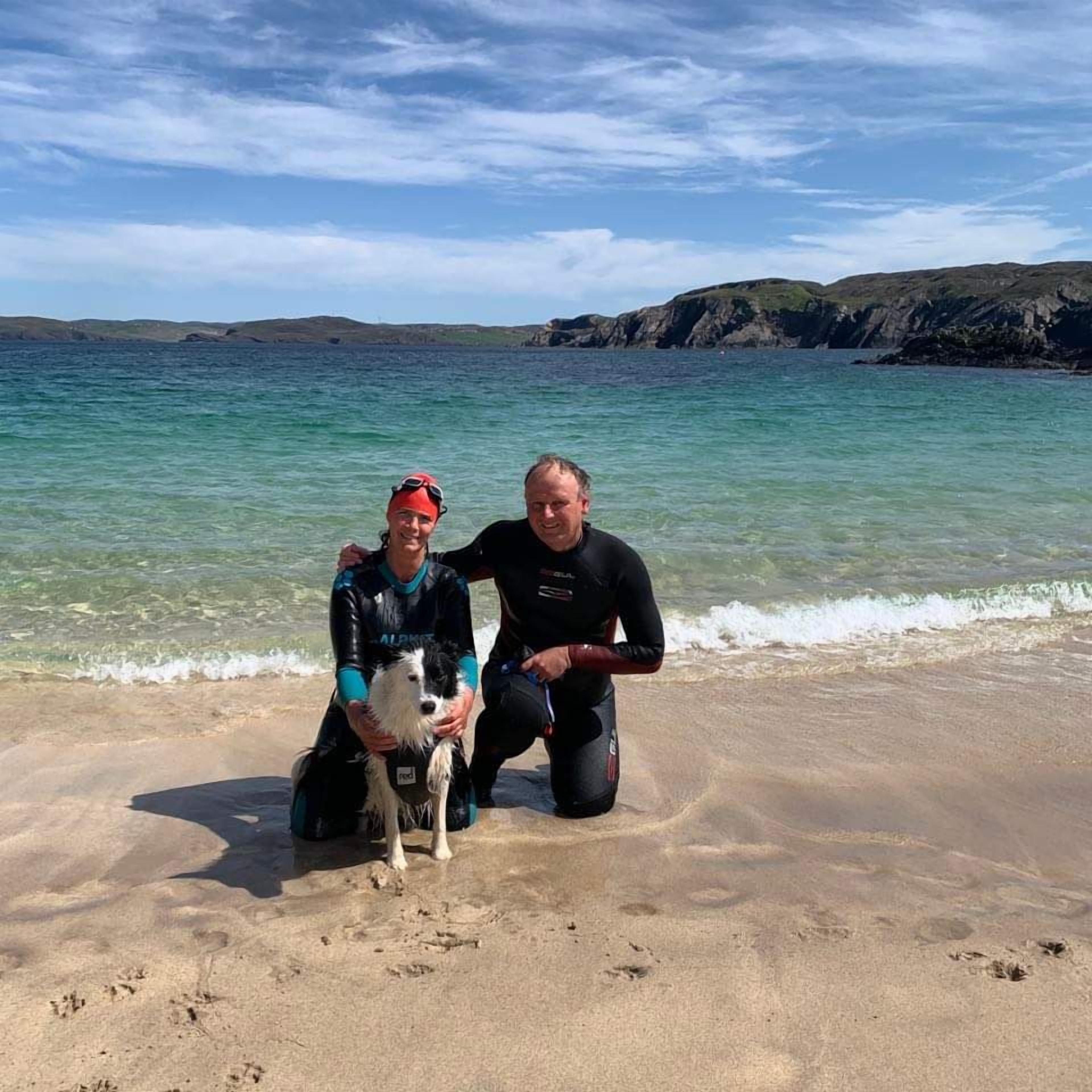 Two people on a beach with a dog, near the water.