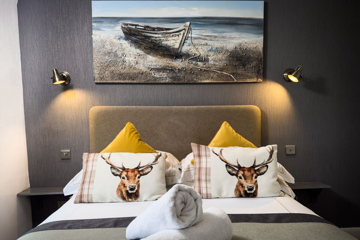 A hotel room with a bed, two nightstands, a painting of a boat on the wall, and two lamps.