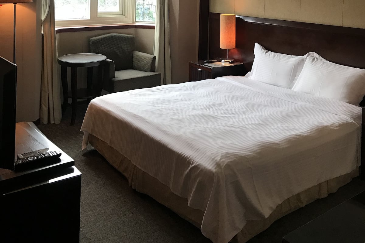 Habitación de hotel con una cama grande, una mesita redonda pequeña, una silla, un televisor y una mesita de noche con lámpara.