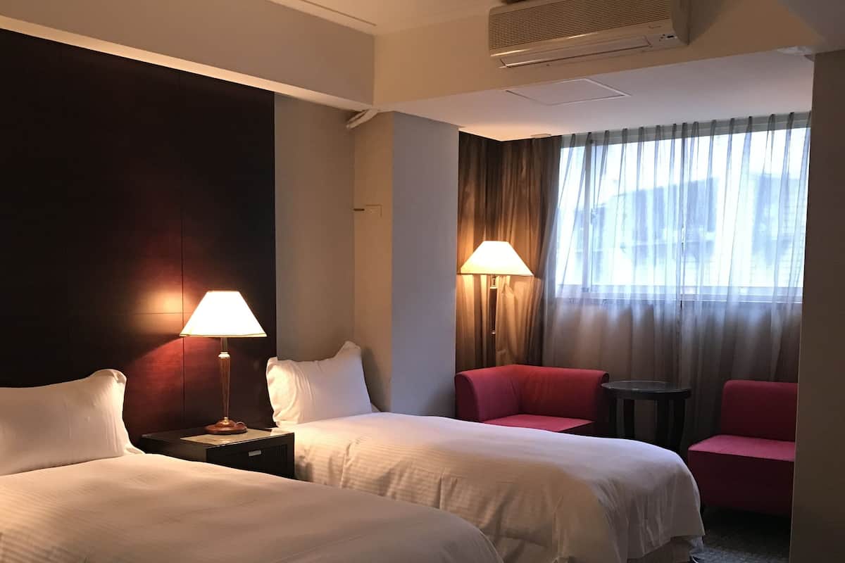 Habitación de hotel con dos camas, un escritorio, una silla, una lámpara y una ventana con cortinas.