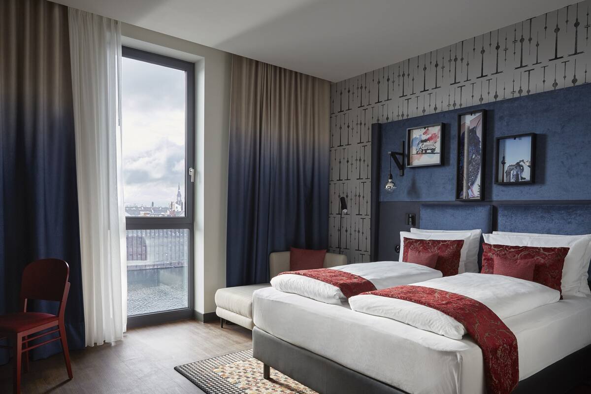 A modern hotel room with a large bed, a chair, and a view of the city through a large window.
