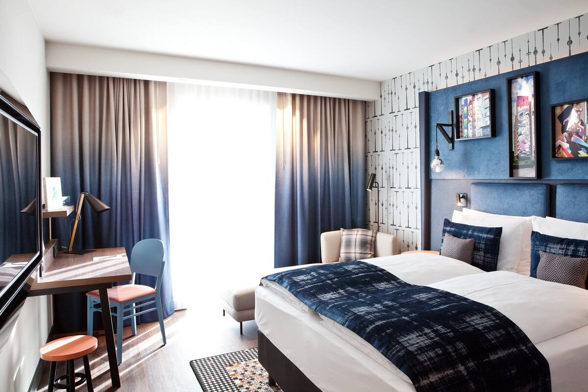 A hotel room with a large bed, a desk with a chair, a small stool, a TV, and a window with curtains.