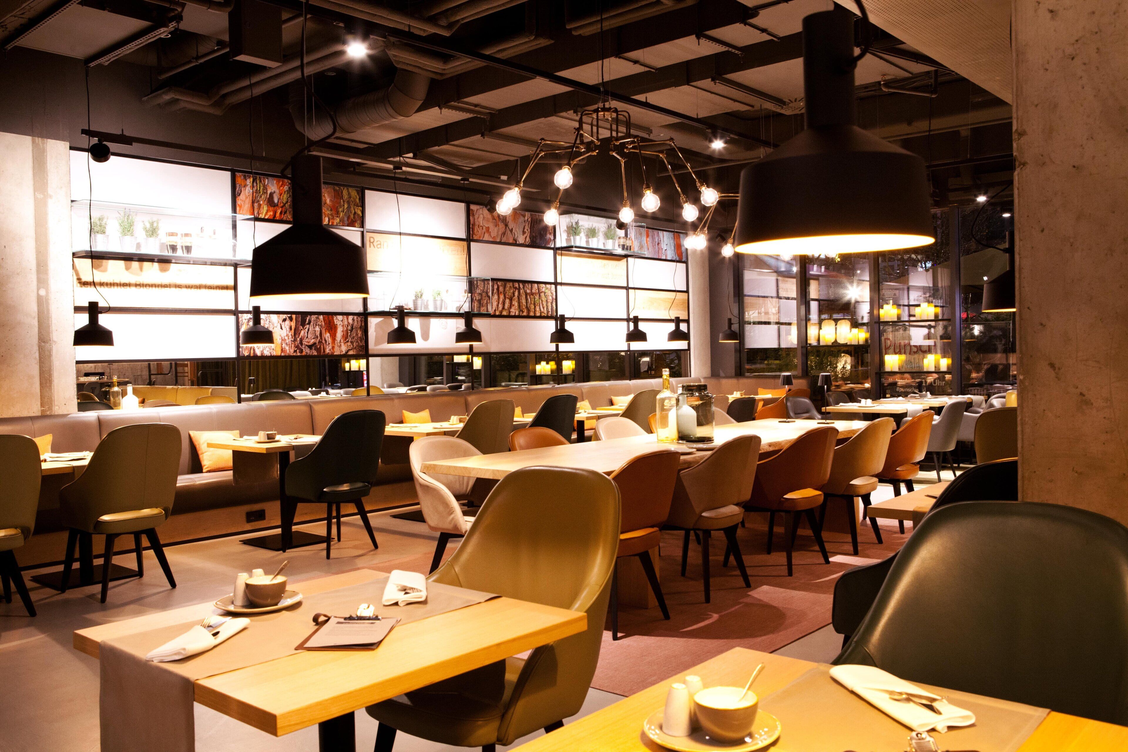 A restaurant interior with multiple tables and chairs, pendant lighting, and a bar area.