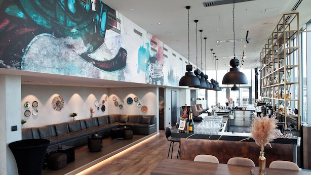 A modern bar area with a long counter, seating, and a large mural on the wall. - Hotel Indigo Berlin - East Side Gallery by IHG (Berlin)