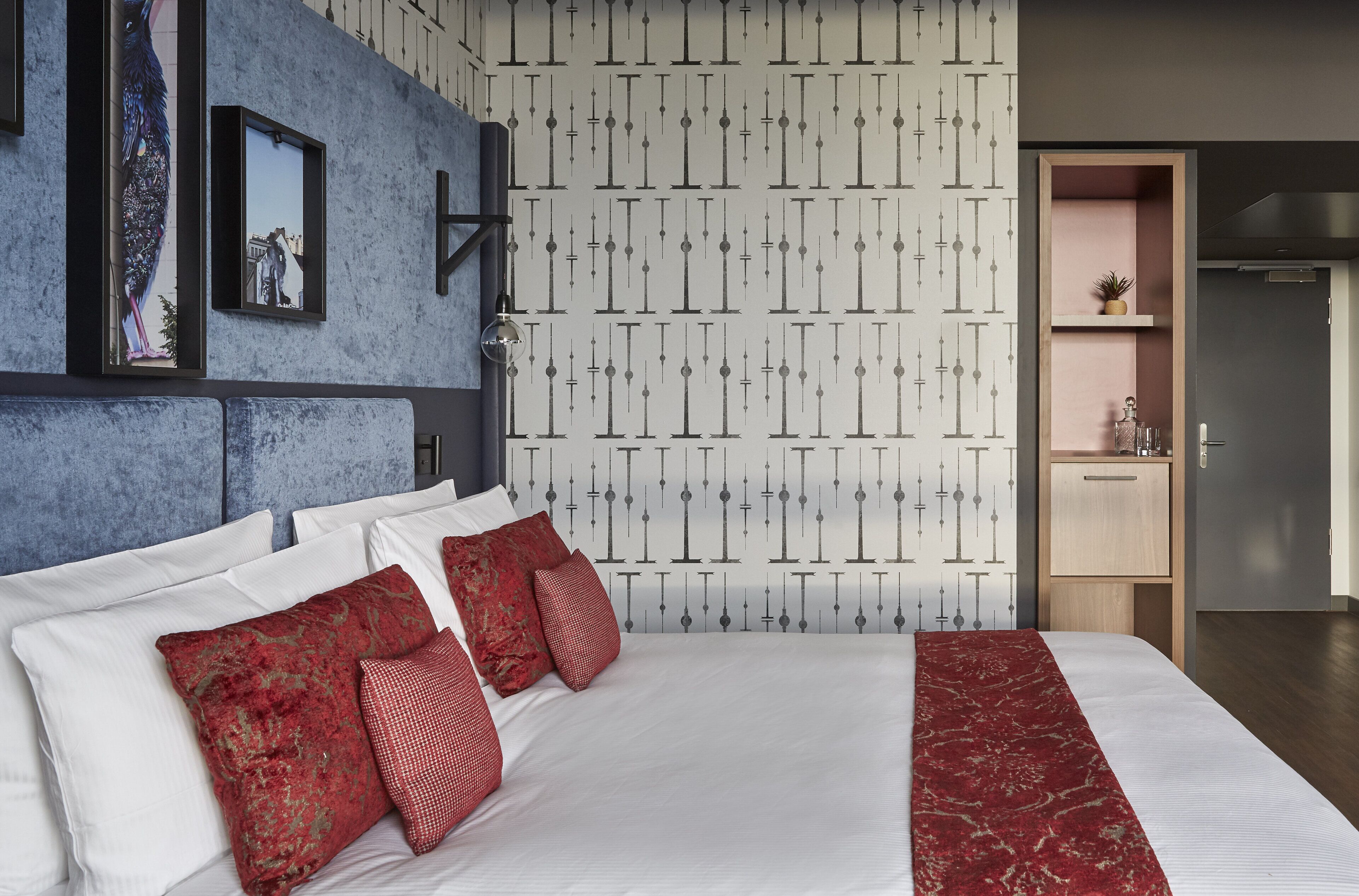 Hotelkamer met een groot bed, rood-witte beddengoed, een blauwe accentmuur, behang met een patroon, en een klein bureau met een plantje.