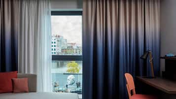 A hotel room with a bed, a desk, a chair, and a view of buildings through a window.