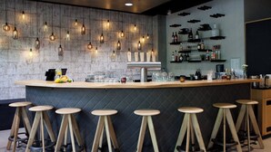 A bar area with a wooden counter, stools, hanging lights, and shelves with bottles and glasses. - Hotel Indigo Berlin - East Side Gallery by IHG (Berlin)