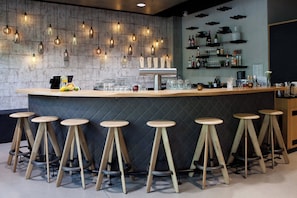 Una zona de bar con mostrador de madera, taburetes, luces colgantes y estantes con botellas y vasos.