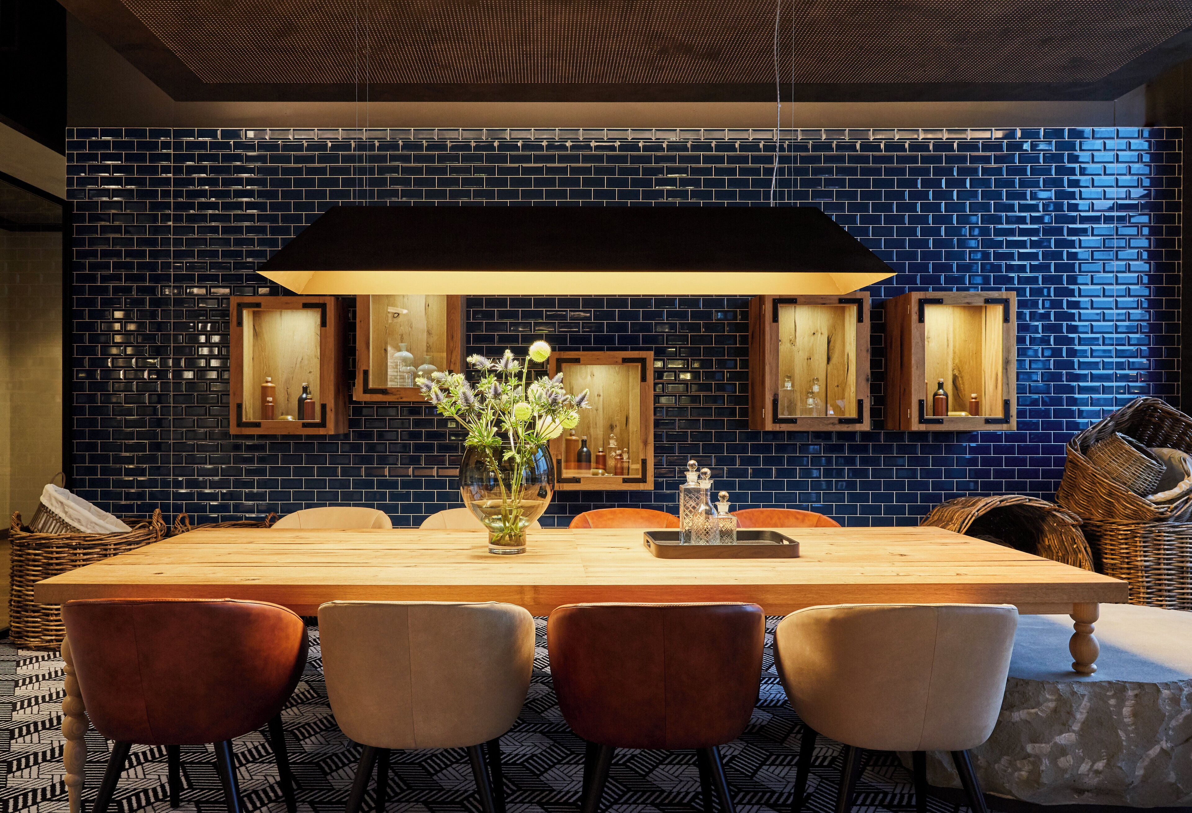 Un comedor con una larga mesa de madera, cuatro sillas, un florero con flores y una pared con azulejos azules y cuadros enmarcados.