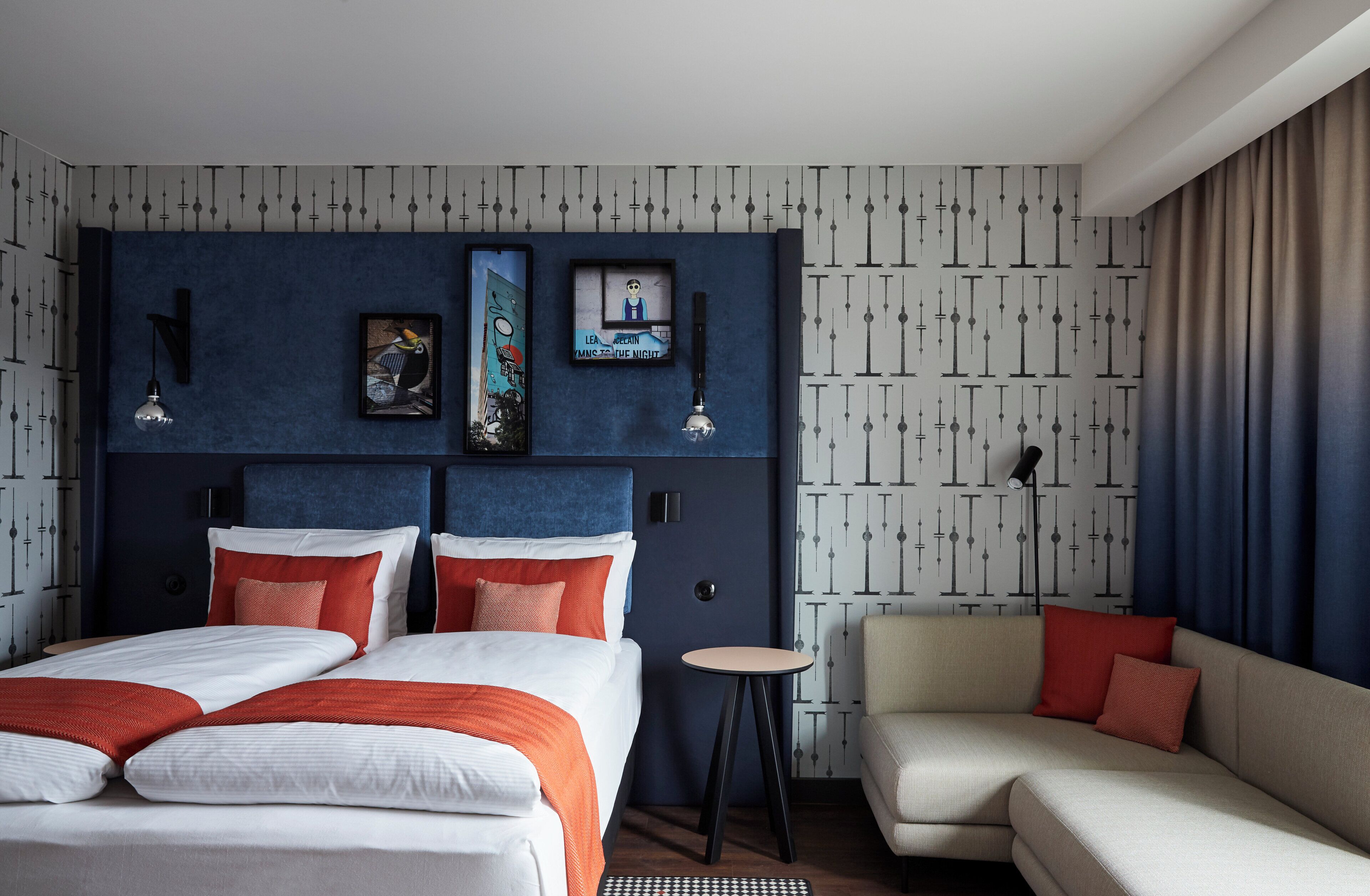 Ein Hotelzimmer mit einem großen Bett, einer Couch und einem kleinen Tisch. An den Wänden befinden sich Kunstwerke und die Tapete ist gemustert.