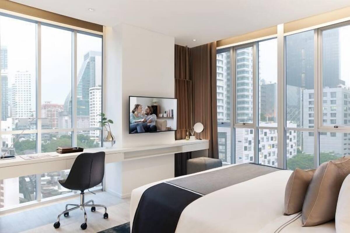 Ein modernes Hotelzimmer mit einem großen Bett, einem Schreibtisch mit Computer, einem Fernseher und Blick auf Stadtgebäude.