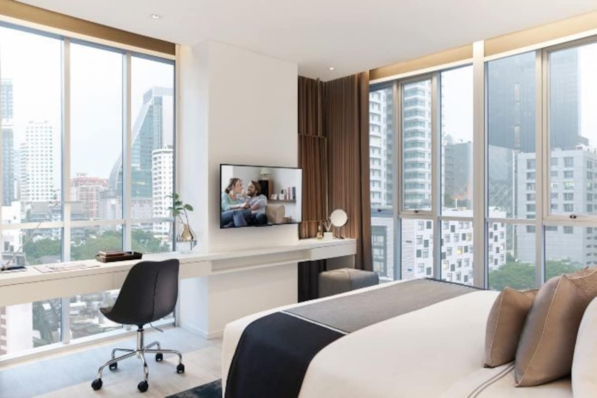 A modern hotel room with a large bed, a desk with a computer, a TV, and a view of city buildings.
