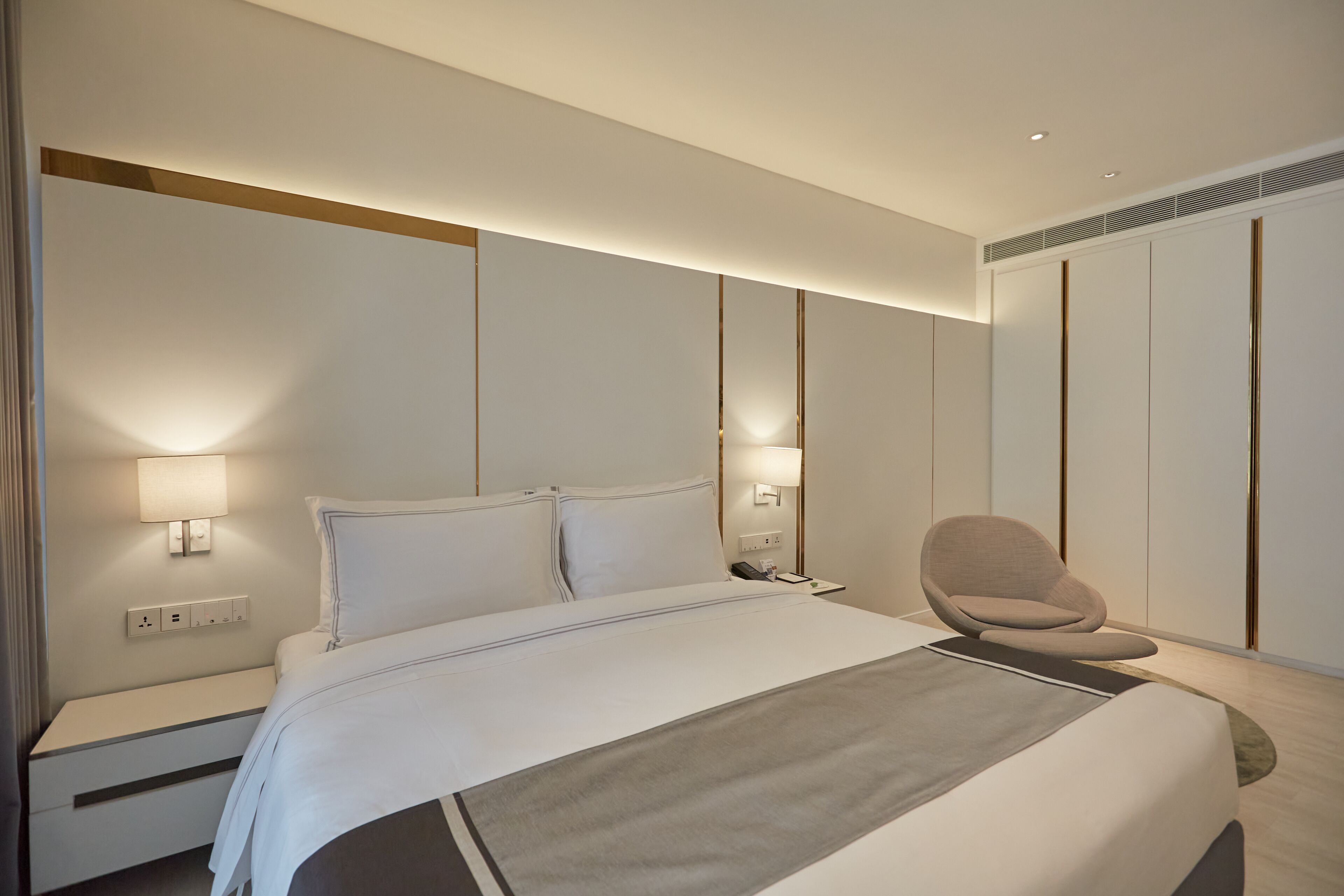 A modern hotel room with a large bed, bedside lamps, a chair, and a wardrobe.
