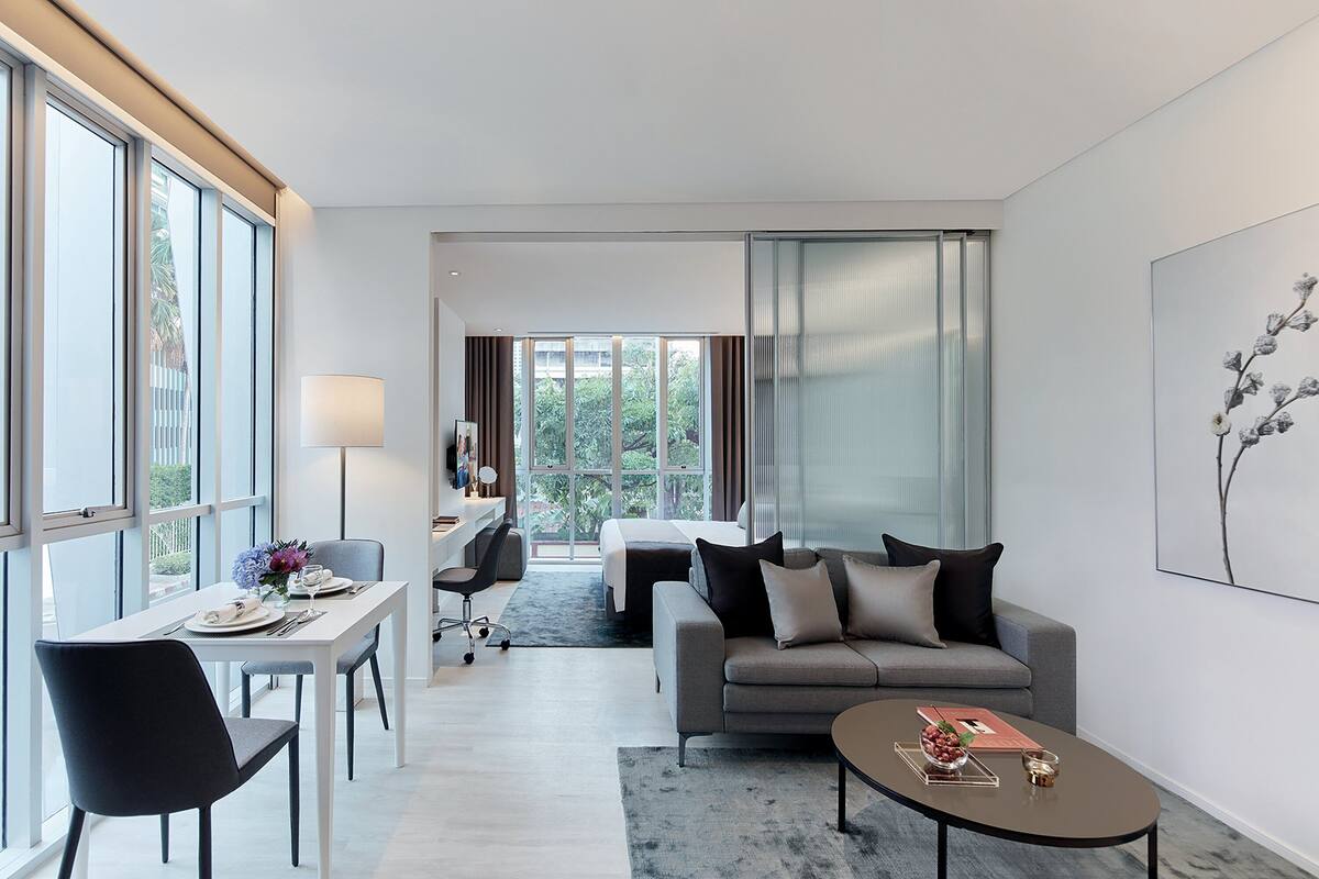 Ein modernes Hotelzimmer mit Essbereich, einer Couch, einem Couchtisch und einem großen Fenster mit Vorhängen.