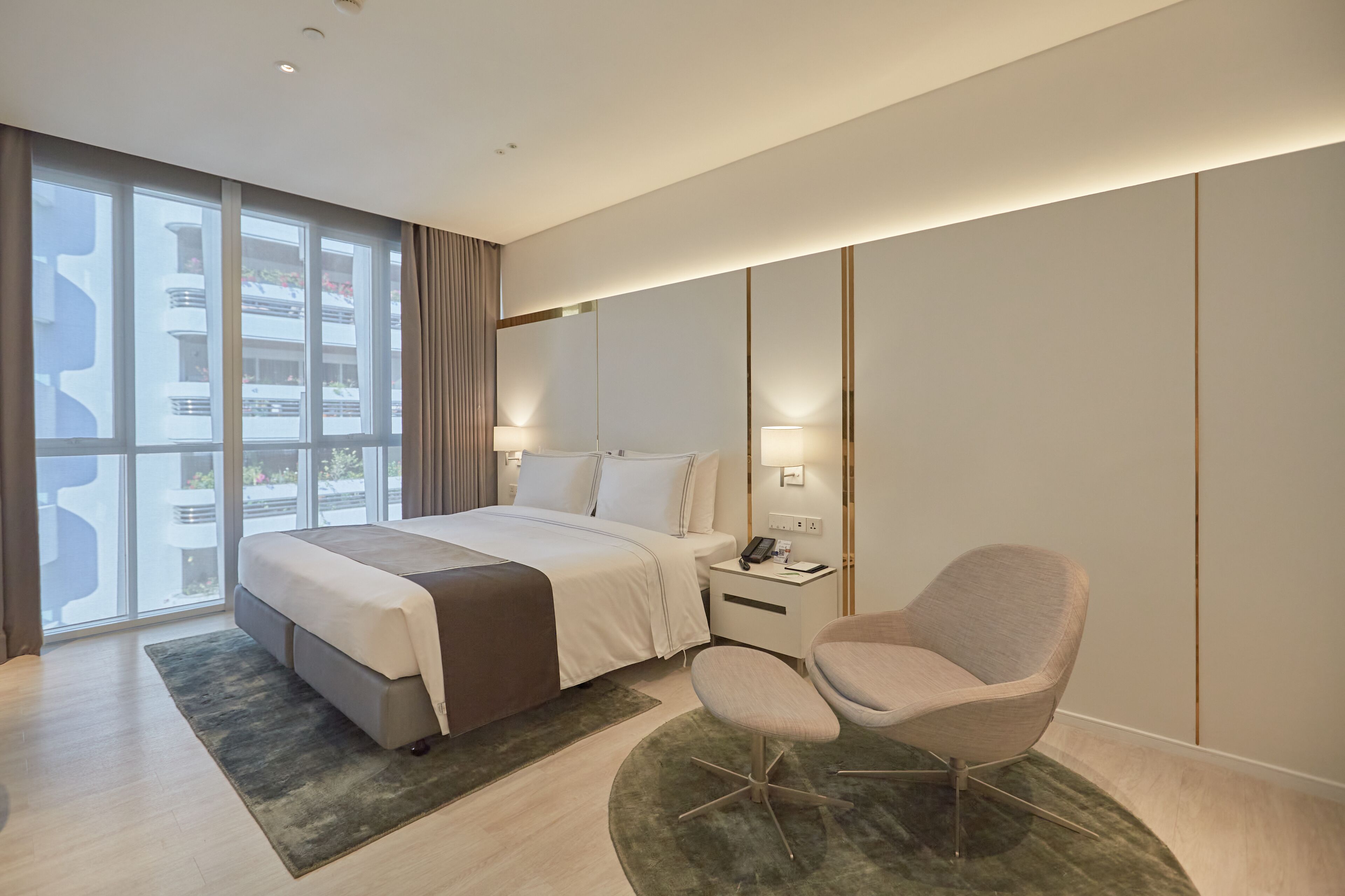 A modern hotel room with a large bed, a sitting area, a nightstand, and a view of the outside.
