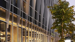 Een terras met tafels en banken, een modern gebouw met een opvallende gevel, en een boom op de voorgrond.