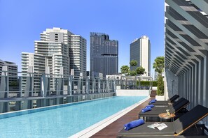 Ein Pool auf dem Dach mit Liegestühlen und einem kleinen Tisch, umgeben von Hochhäusern.