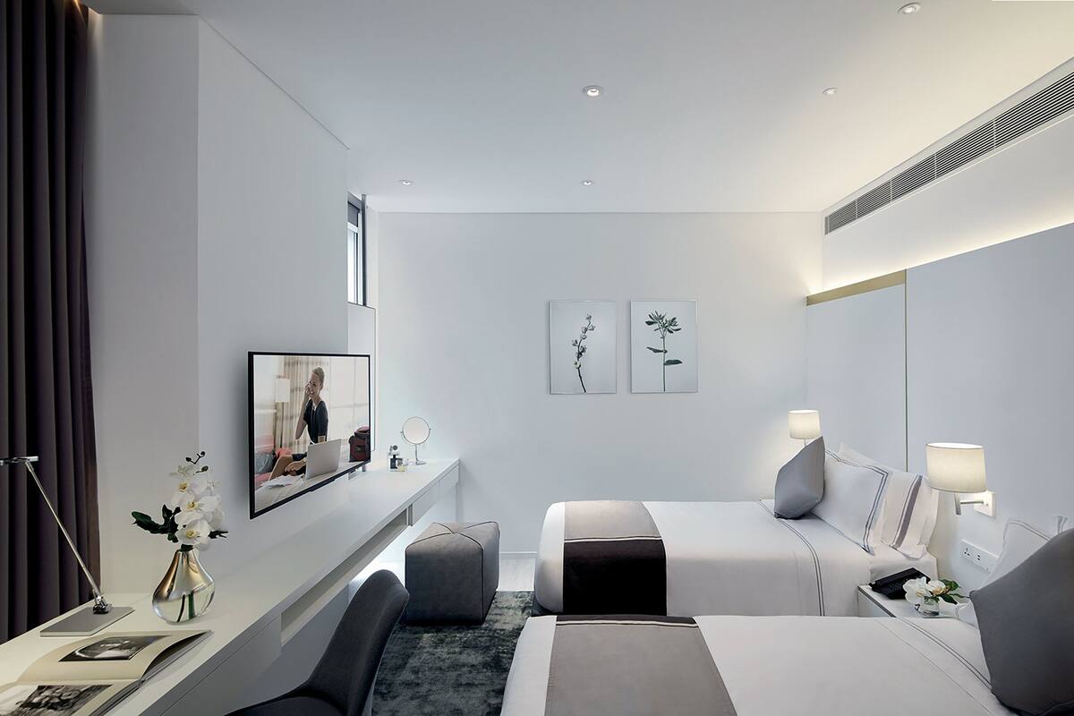 Ein modernes Hotelzimmer mit Bett, Schreibtisch, Stuhl, einem kleinen Tisch und einem Spiegel. Zwei Bilderrahmen sind an der Wand angebracht, und es gibt ein Fenster mit Vorhängen.