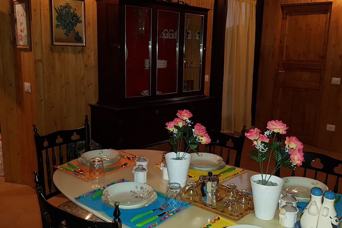 A dining area with a wooden table set for four, surrounded by chairs. A cabinet with glass doors is visible in the background.
