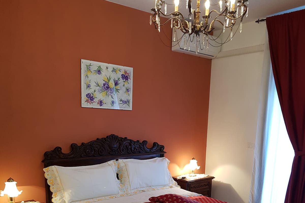 A bedroom with a large bed, a chandelier, a nightstand, a lamp, a rug, and a painting on the wall.