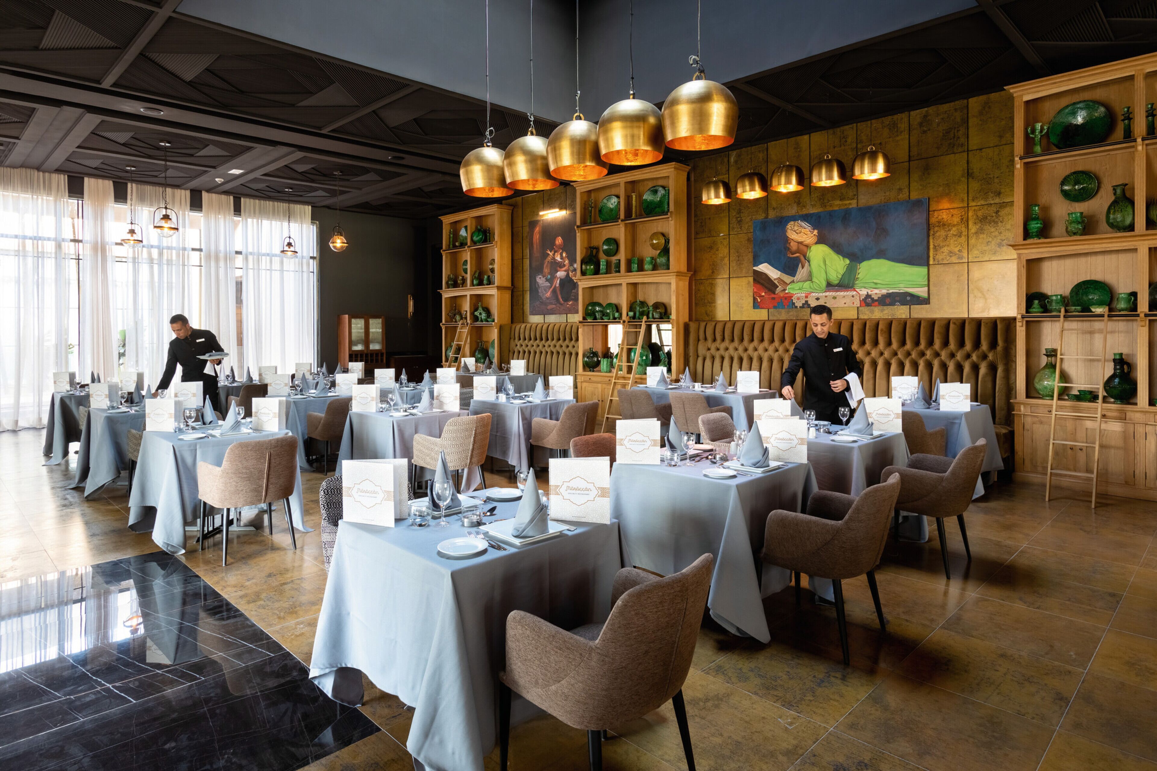 Un restaurante con varias mesas dispuestas para comer, un barman preparando bebidas y un gran mural en la pared.