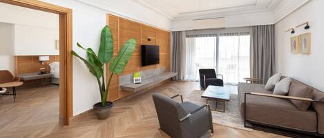 Una habitación de hotel moderna con suelo de espiga, un televisor de pantalla plana montado en una pared de paneles de madera, una zona de estar con sofá y sillones, y una planta grande en maceta.