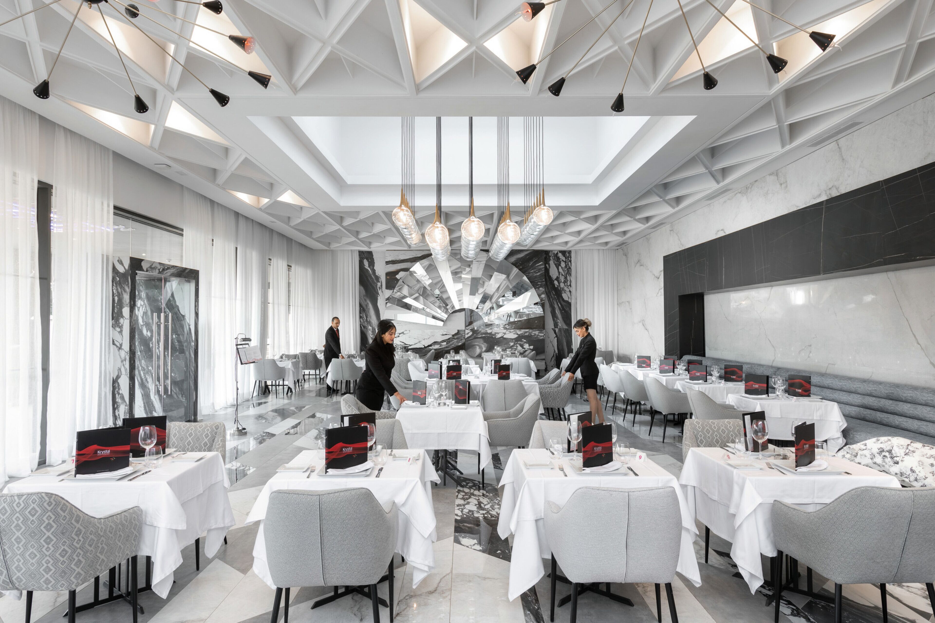 Un comedor moderno con mesas dispuestas para una comida, un mural grande en la pared y un techo con patrones geométricos y luces colgantes.