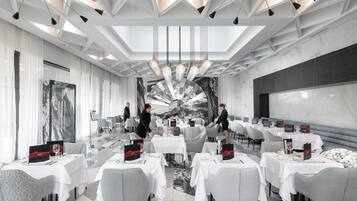 Un comedor moderno con mesas dispuestas para una comida, un mural grande en la pared y un techo con patrones geométricos y luces colgantes.