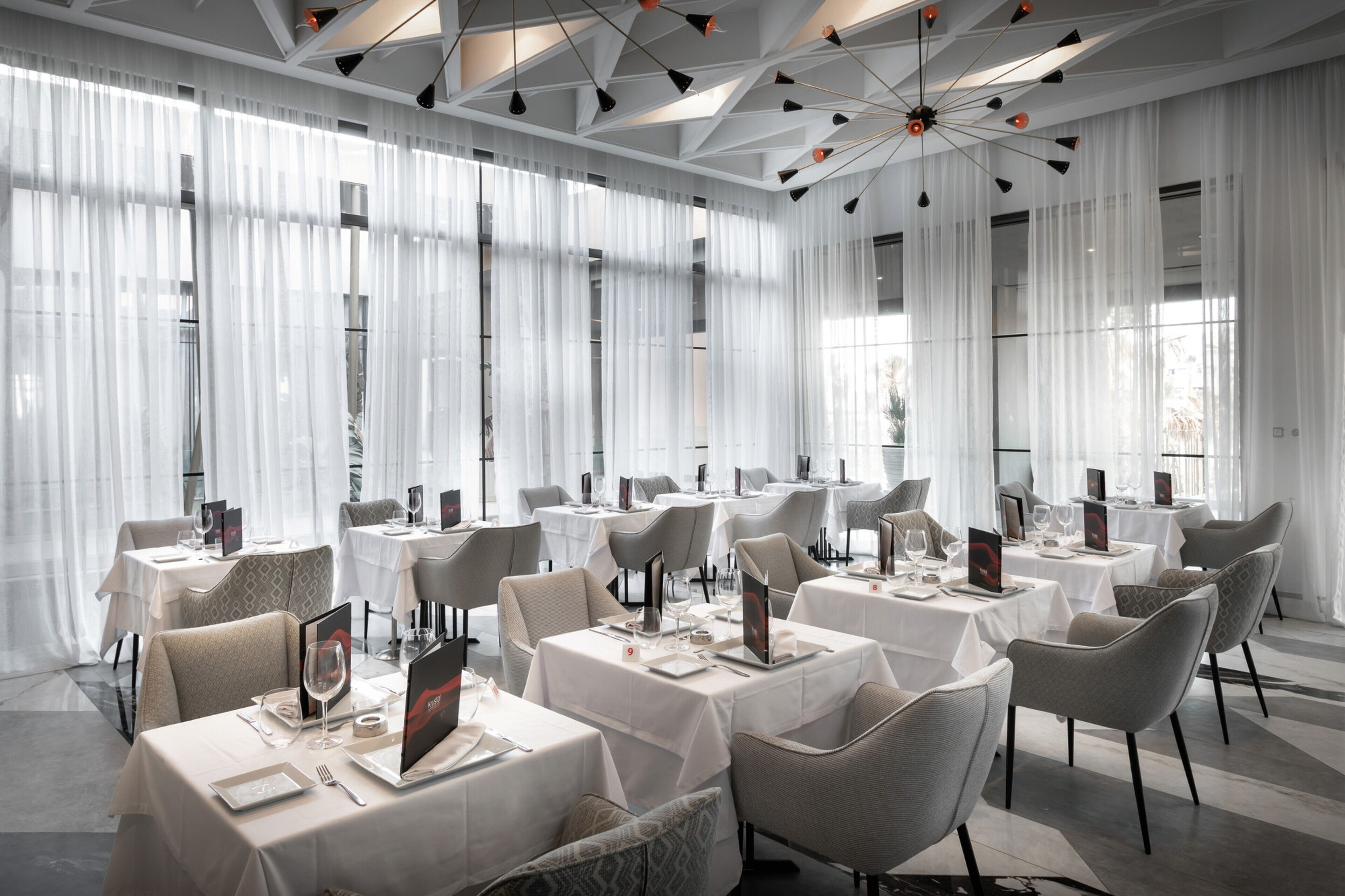 A modern dining area with white tables and chairs, large windows with sheer curtains, and a geometric ceiling design.