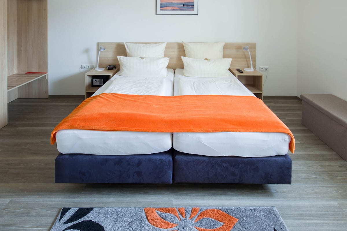 A double bed with an orange blanket, two bedside tables with lamps, a wooden wardrobe, and a framed picture on the wall.