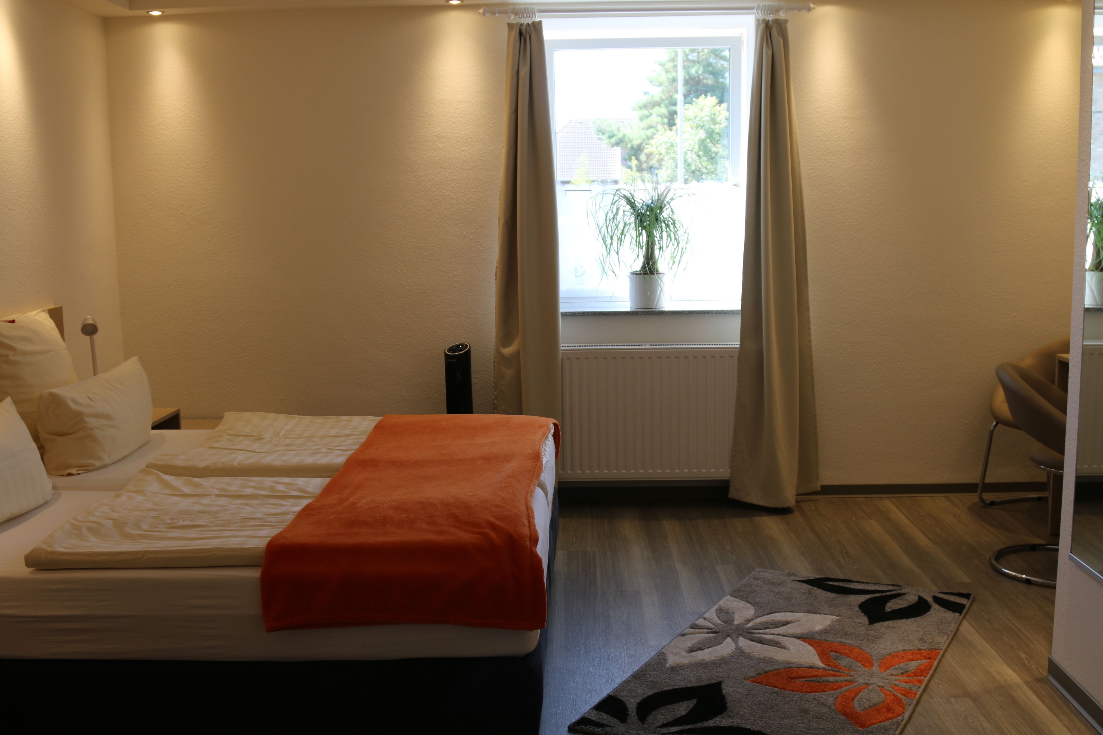 A hotel room with two beds, a window with curtains, a radiator, a potted plant, and a chair.