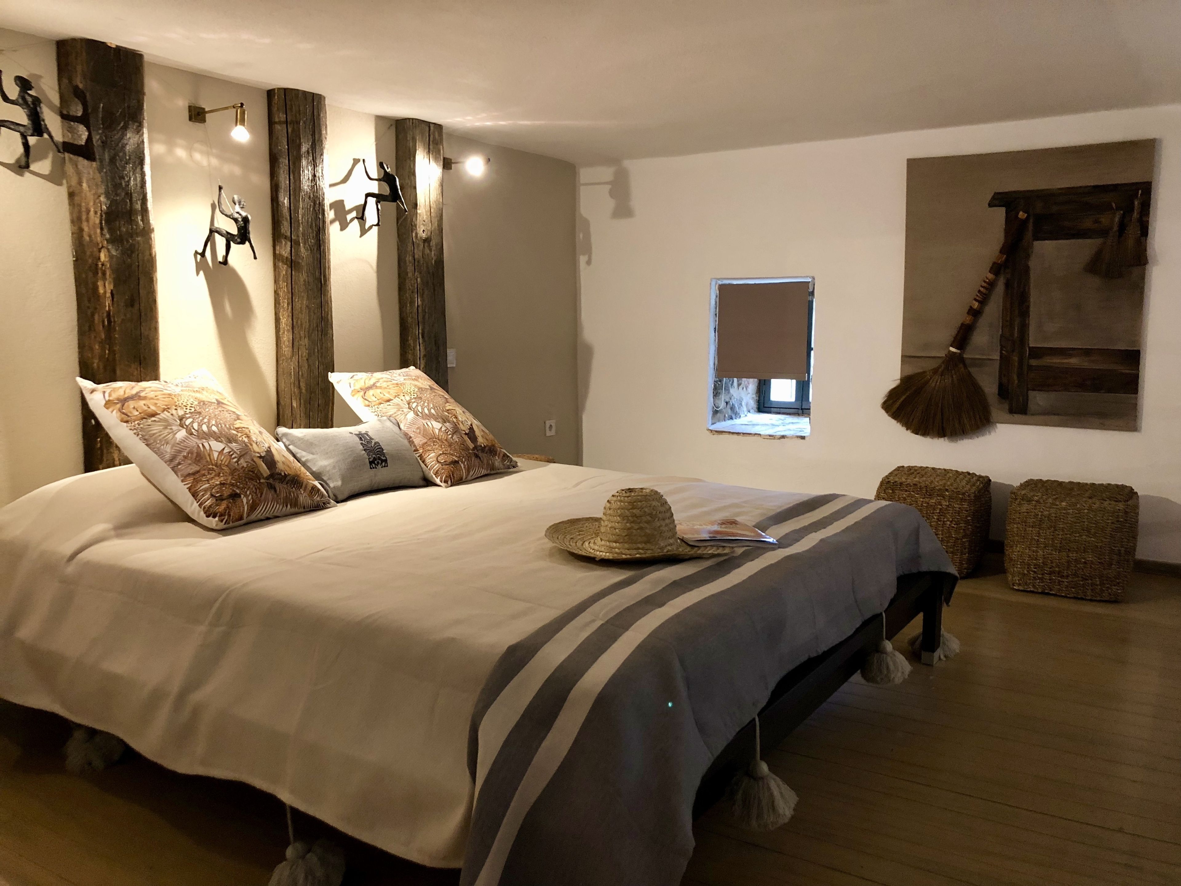 Une chambre avec un grand lit, des poutres en bois, une fenêtre et un balai accroché au mur.