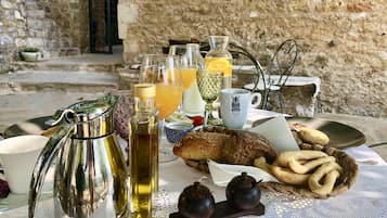 A breakfast setup with a coffee pot, bottles, glasses, and bread on a table outdoors with stone walls in the background.