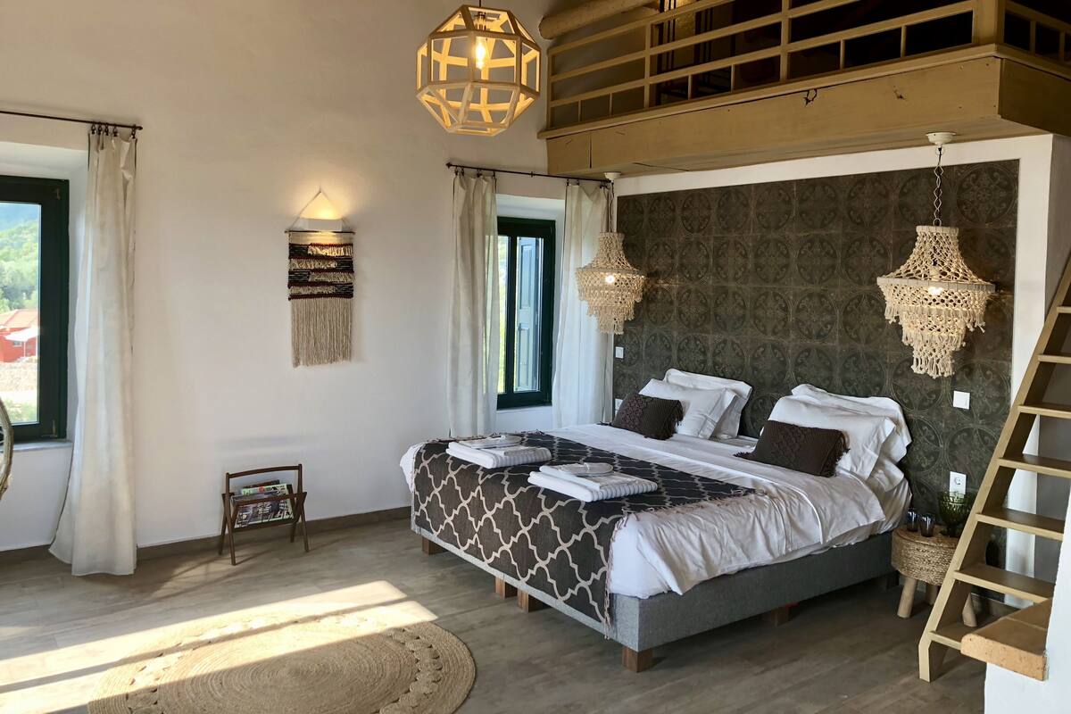 A bedroom with a large bed, two hanging light fixtures, a wooden ladder, and a window with curtains.