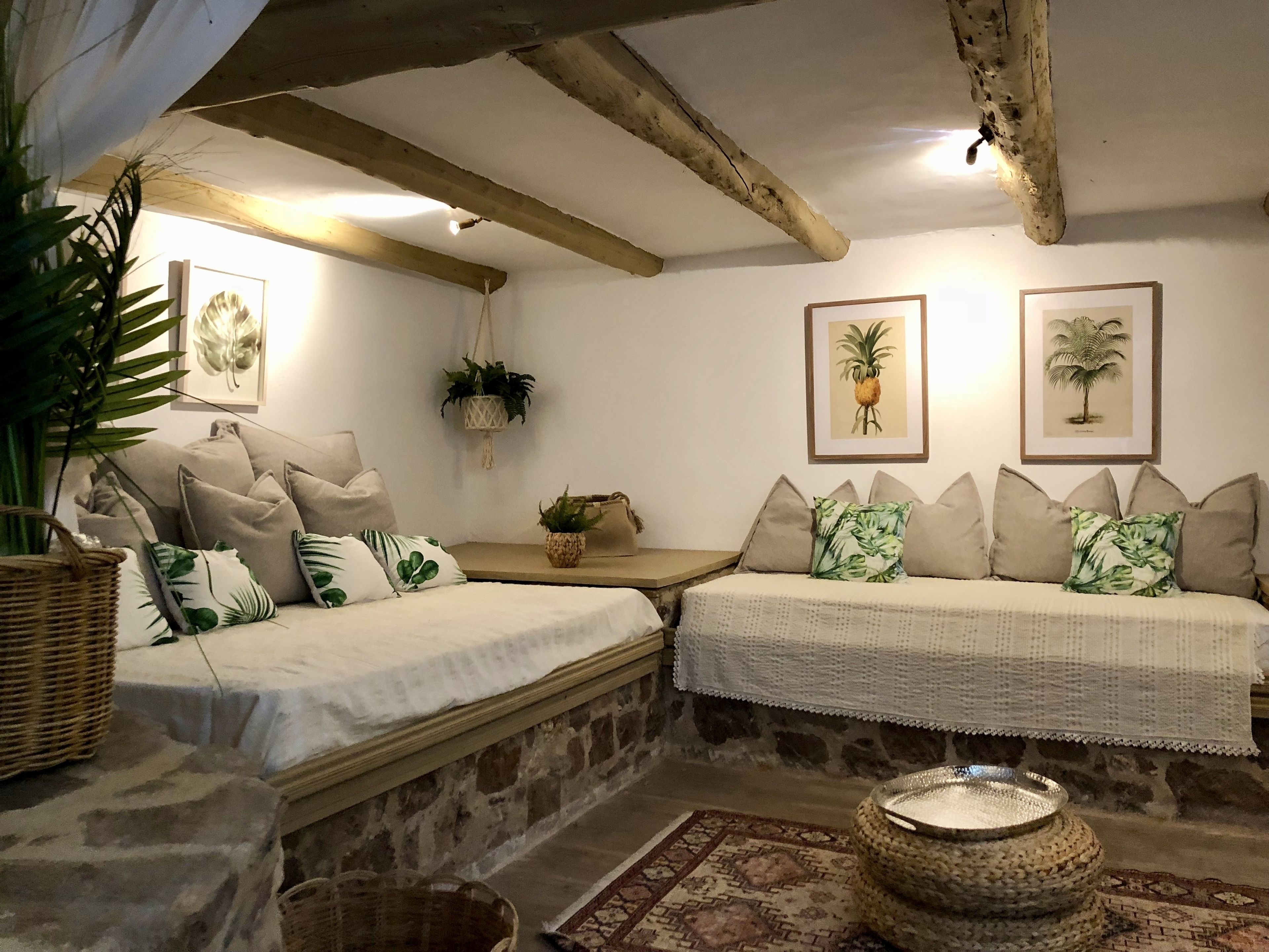 A cozy room with a stone wall, two beds with matching pillows, a woven basket, a woven stool, and framed botanical prints on the wall.