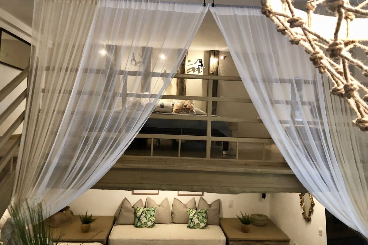 A cozy room with a bed, a window with sheer curtains, and a small table with a plant.