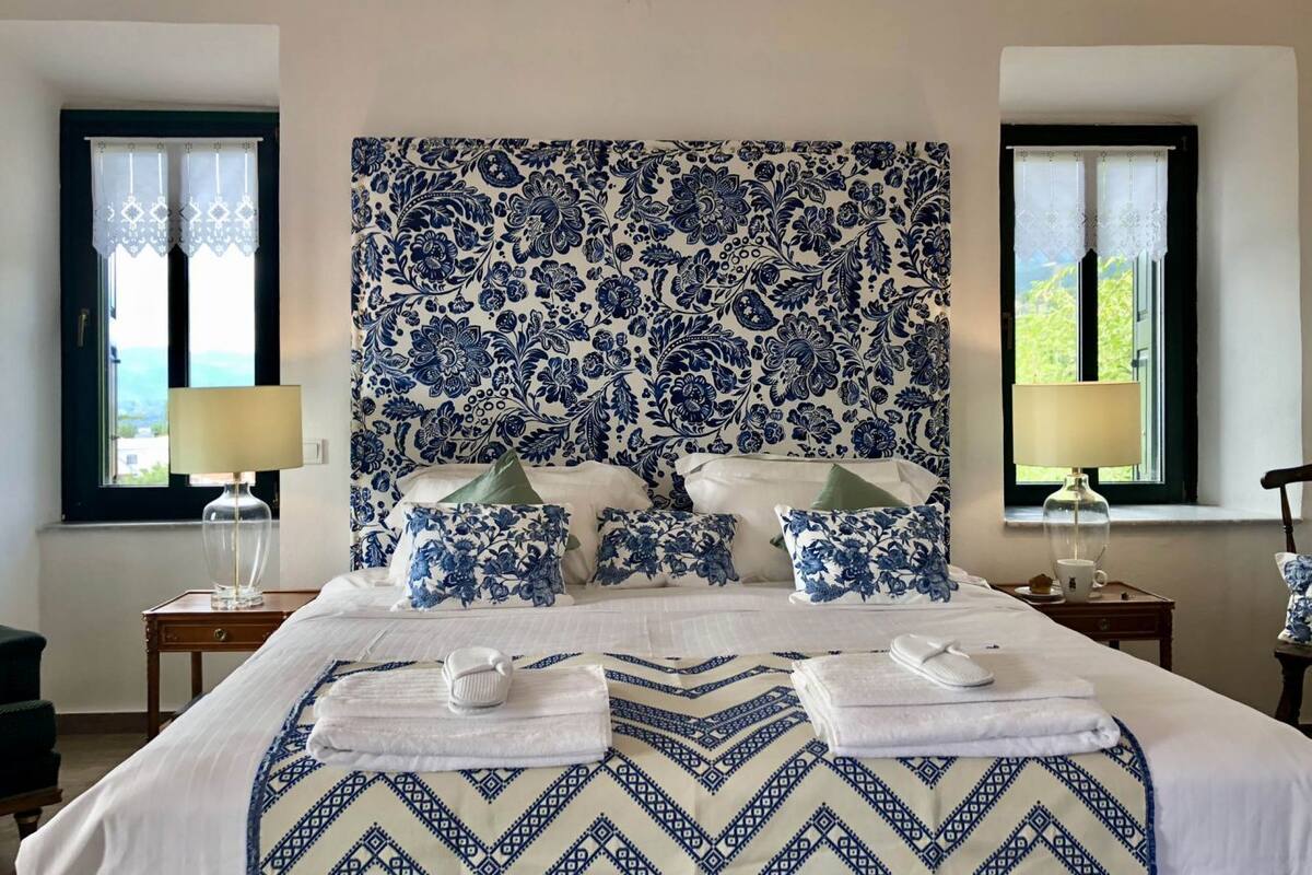 A hotel room with a large bed, floral-patterned headboard, two bedside lamps, and a view of the outdoors through windows.