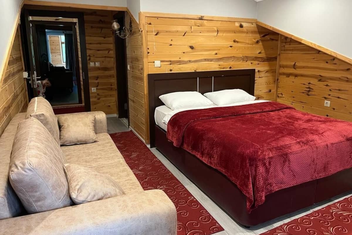A bedroom with a wooden sloped ceiling, a bed with a red blanket, a beige sofa, and a patterned rug.