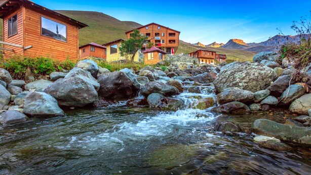 A wooden lodge with a stream flowing through it, surrounded by large rocks and hills in the background. - Yedigoller Hotel & Restaurant (Çaykara)