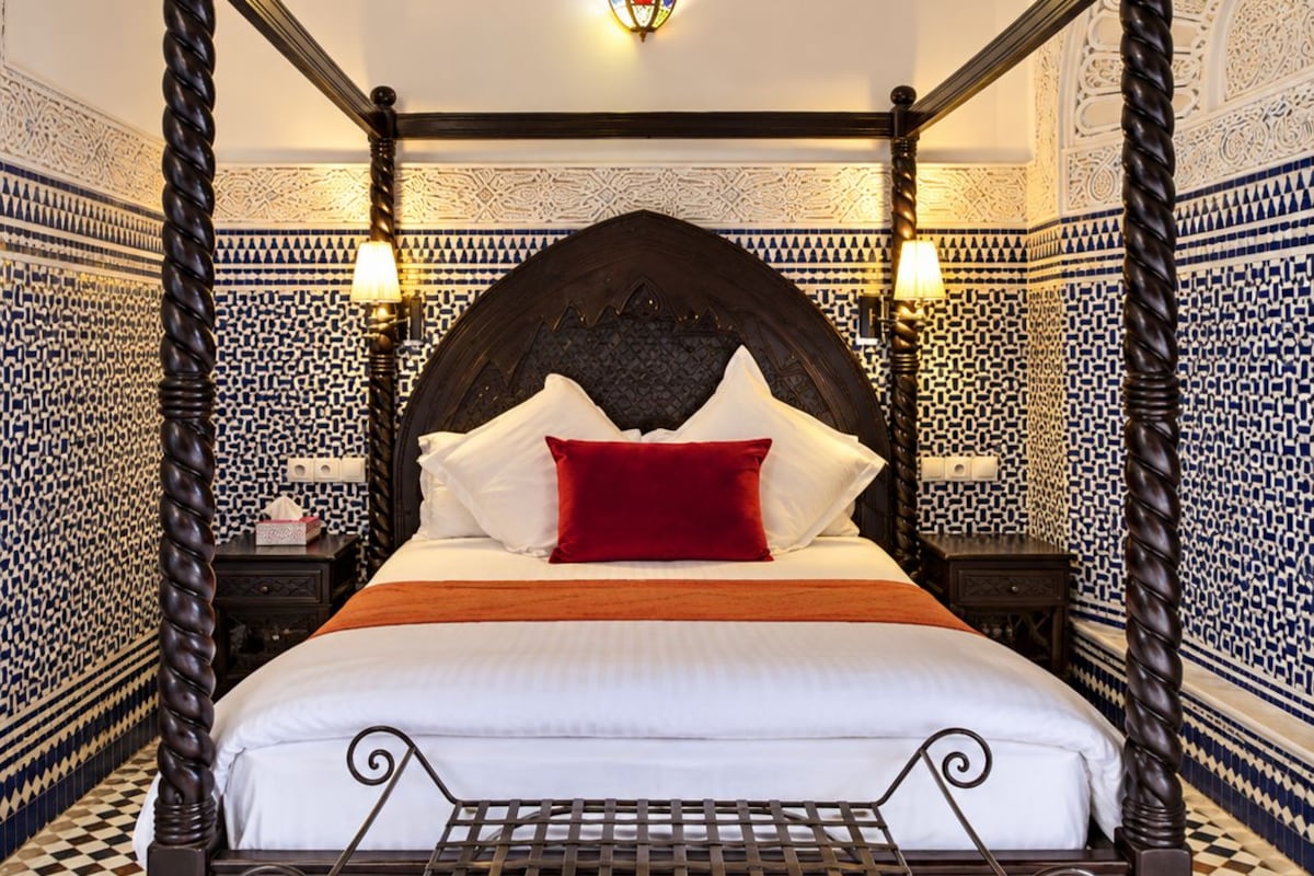 Uma cama com dossel de madeira escura, roupa de cama branca e almofada vermelha de destaque.
