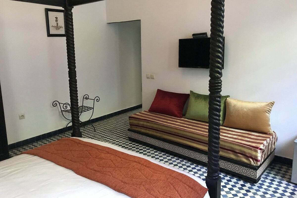 Uma cama com dossel com um cobertor marrom, piso xadrez, um sofá com almofadas listradas e uma TV na parede.