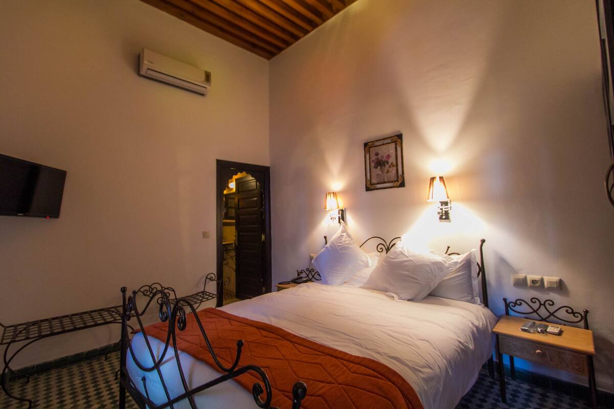 Quarto com cama, mesas de cabeceira, ar condicionado, televisão fixada na parede e uma porta que dá para outro cômodo.