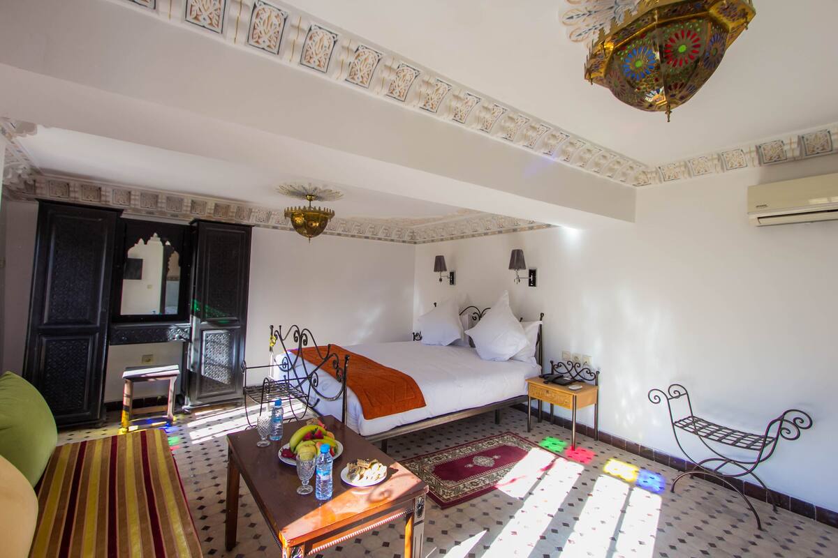 Quarto de hotel com cama, uma pequena mesa com frutas e água, um banco e uma luminária decorativa no teto.