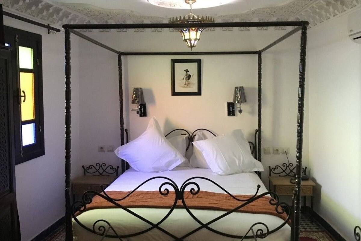 Uma cama com dossel e estrutura de madeira, roupa de cama branca e um cobertor estampado. Há duas luminárias fixadas na parede e um quadro emoldurado acima da cama.