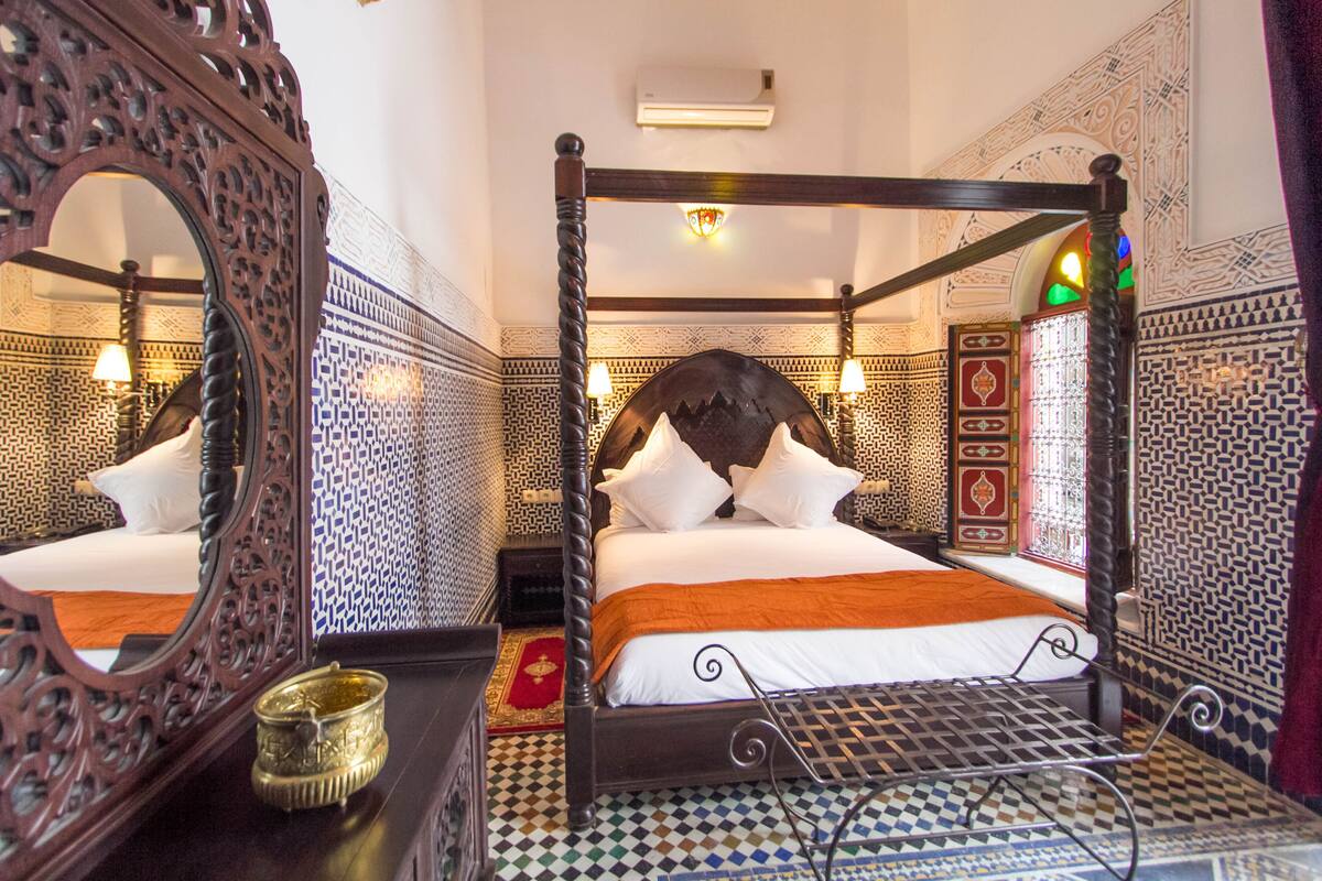 Uma cama de dossel com roupa de cama branca e um cobertor laranja, rodeada por paredes com padrões elaborados e um espelho grande e ornamentado.