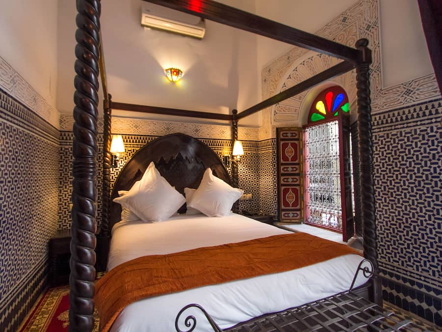Una cama con dosel, una manta marrón, un papel tapiz estampado y una ventana de vitral.